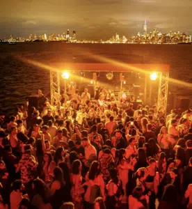 A large crowd dances at an outdoor night party on a waterfront, energized by New York music, with city skyline lights and tall buildings visible across the water.